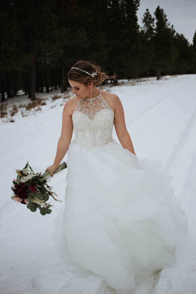 Bride in white lace shoulder less dress holding blush pink bouquet walking in snowy field 