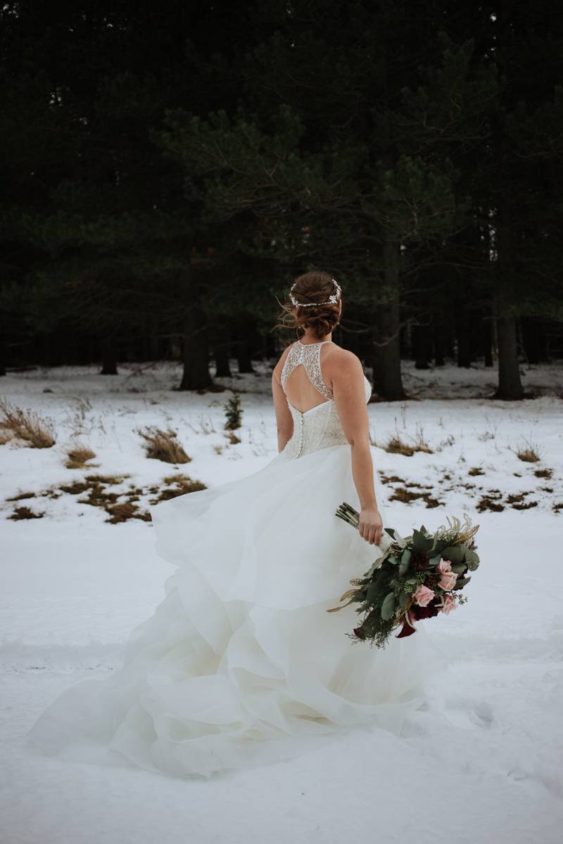 Bride in white open back dress holding blush pink bouquet walking in snowy field 