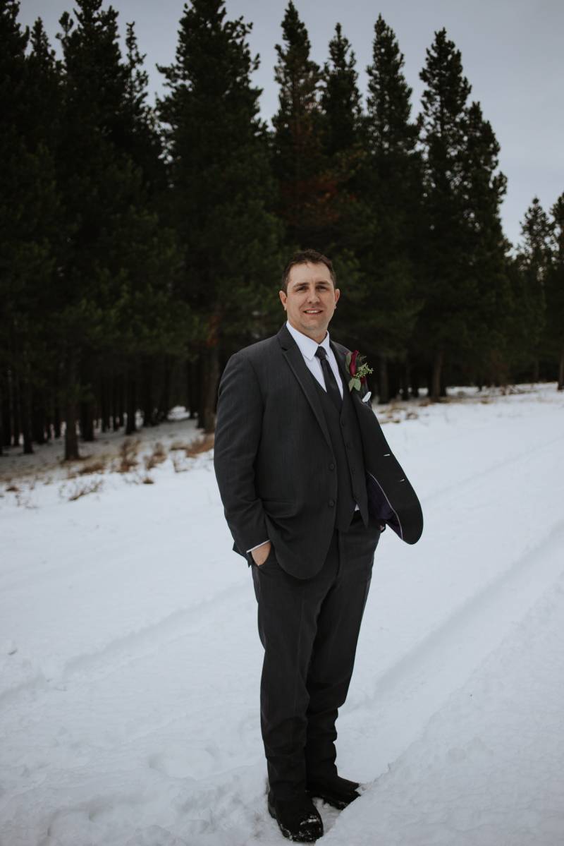 Groom stands on snowy road hands in pockets in front of dense green forest 