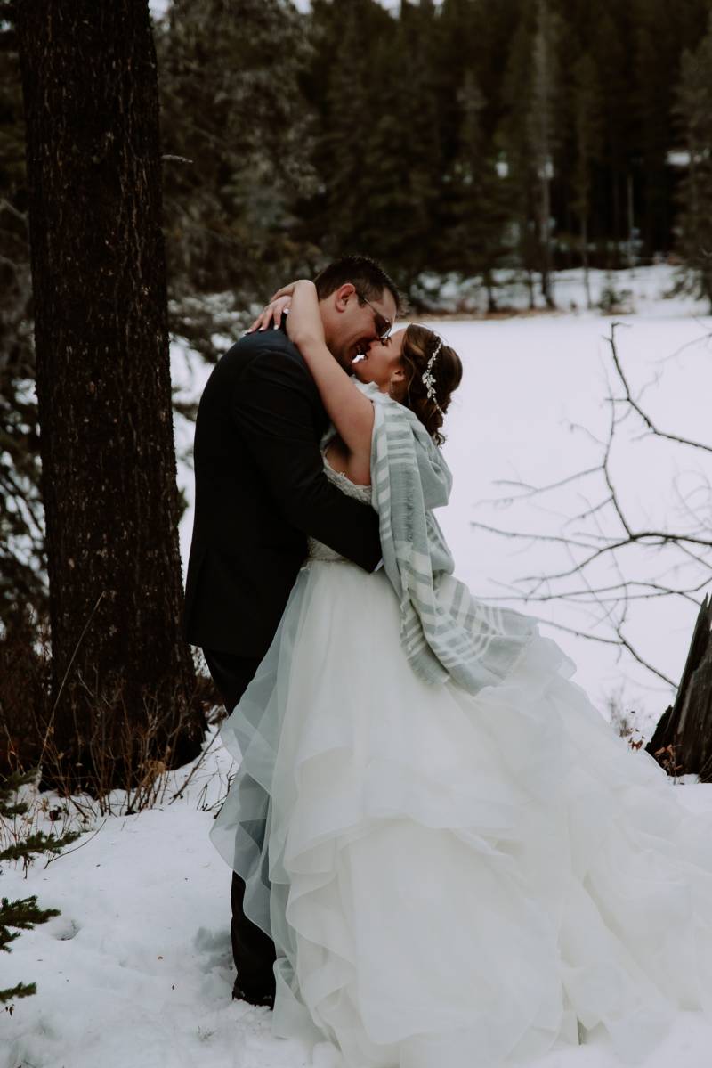 Bride and groom kiss embracing in front of snowy forest field 