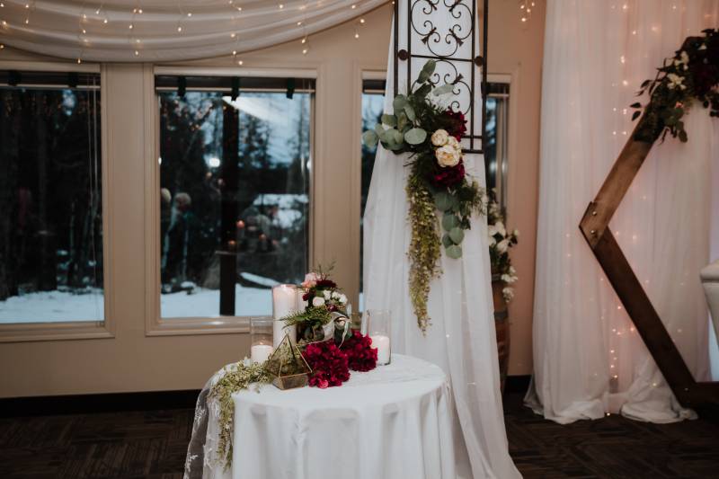 White table with burgundy and white floral arrangements in front of window with fairy lights hanging