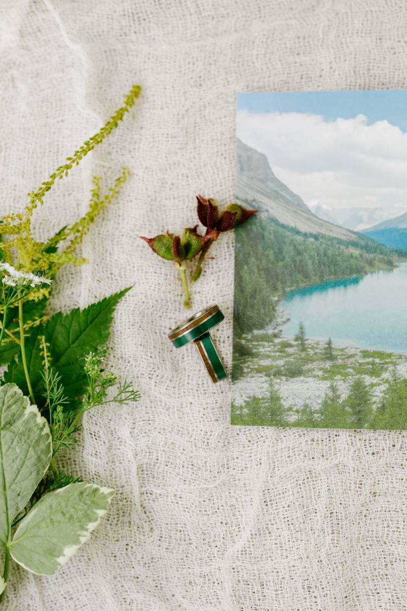 Bronze and green wedding ring flat lay beside image of mountain lake