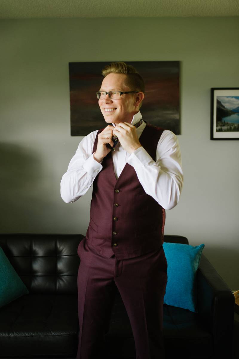Man in burgundy suit adjusts bowtie while smiling