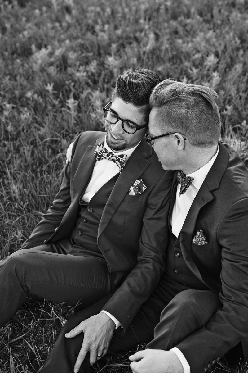 Grooms sitting smiling in grassy field hand on leg
