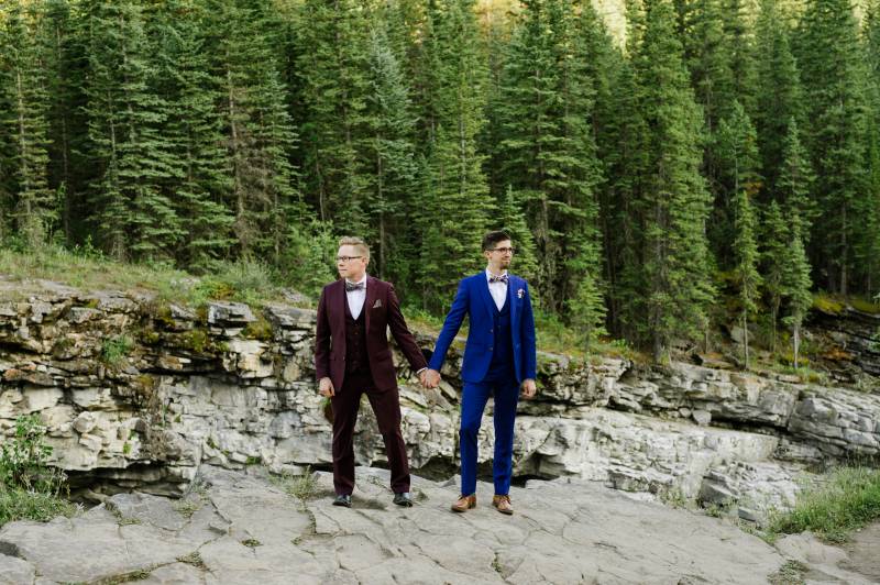 Grooms holding hands standing on rocky forest bank