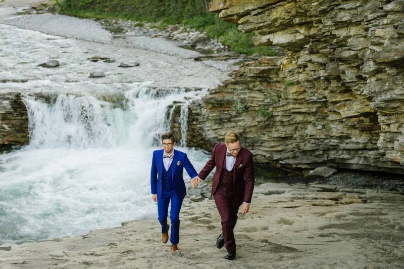 Grooms walk together holding hands up rocky slate riverbank