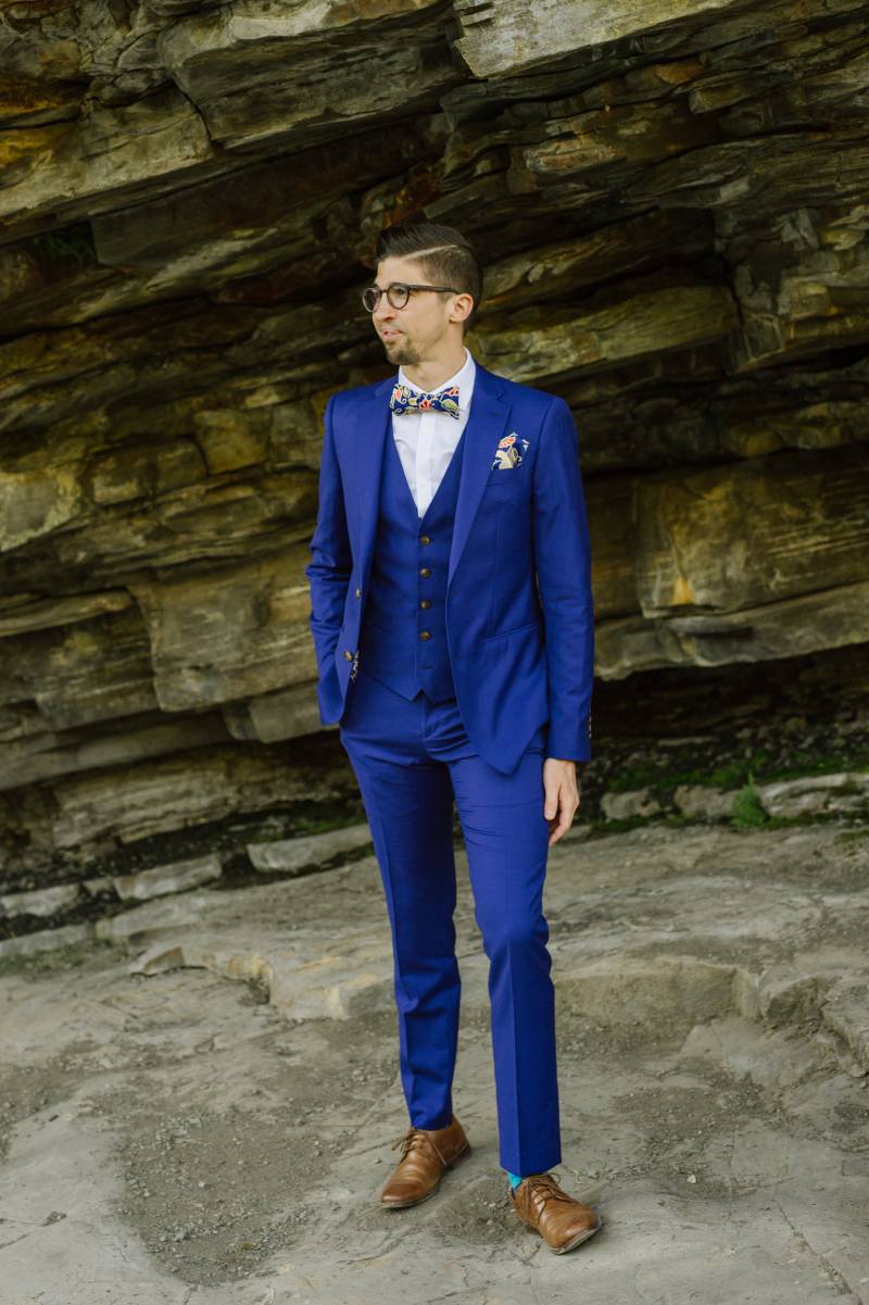 Groom in blue suit stands hand in pocket on rocky slope