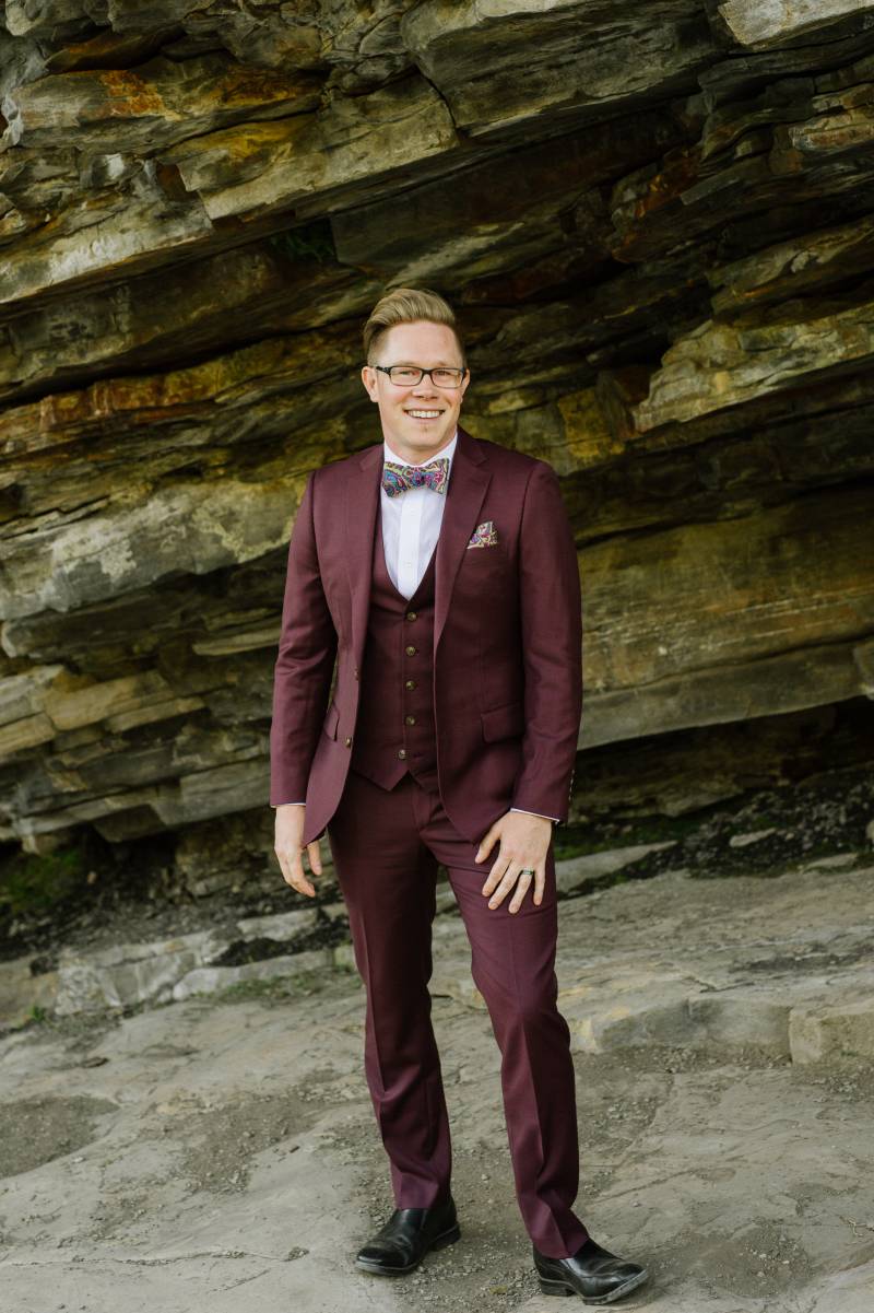 Groom in burgundy suit stands on rocky slate bank
