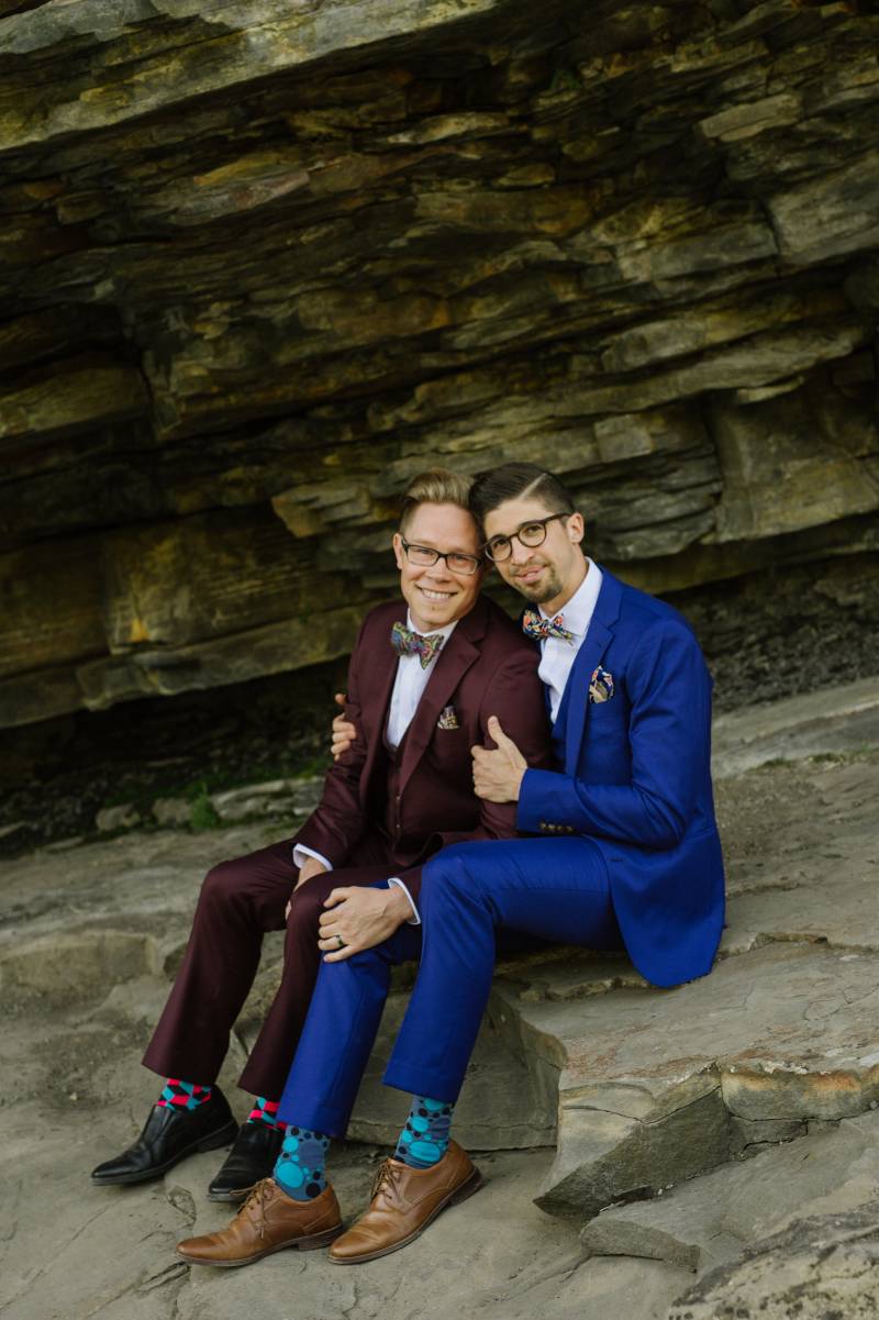 Grooms sit together embraced underneath slate wall