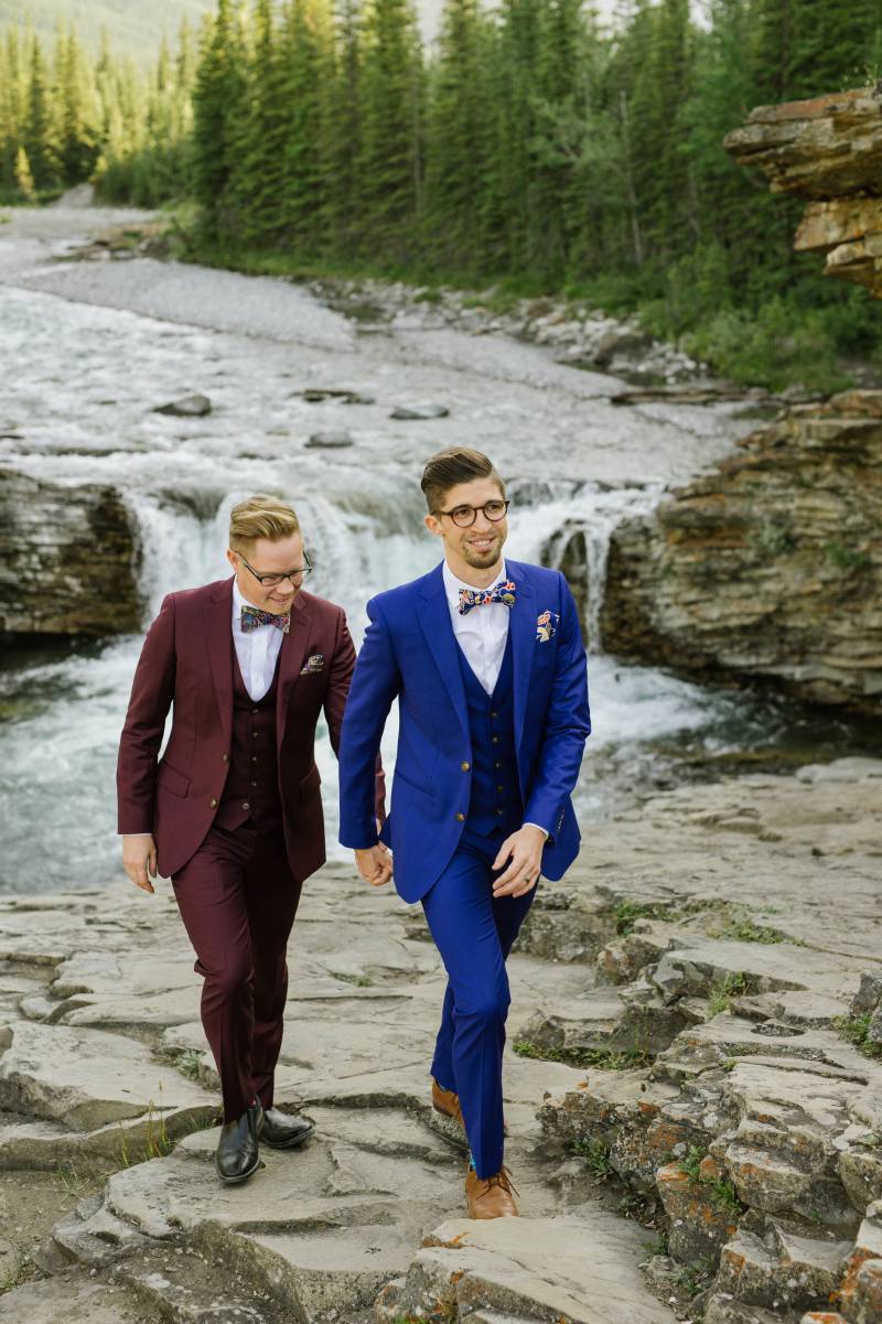 Grooms walk together up rocky riverbank 