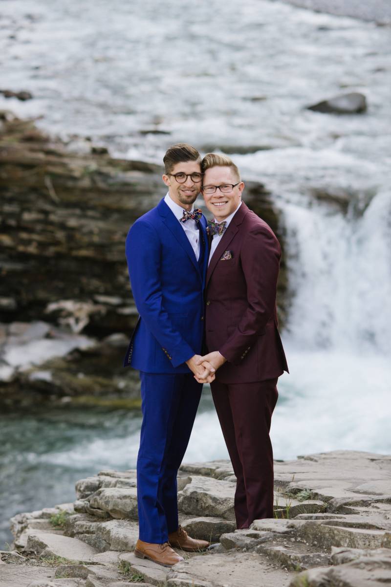 Grooms hold hands in front leaning on one another in front of waterfall