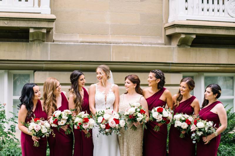 Bride wearing white dress and bridesmaids wearing burgundy dresses stand holding white and burgundy bouquets 