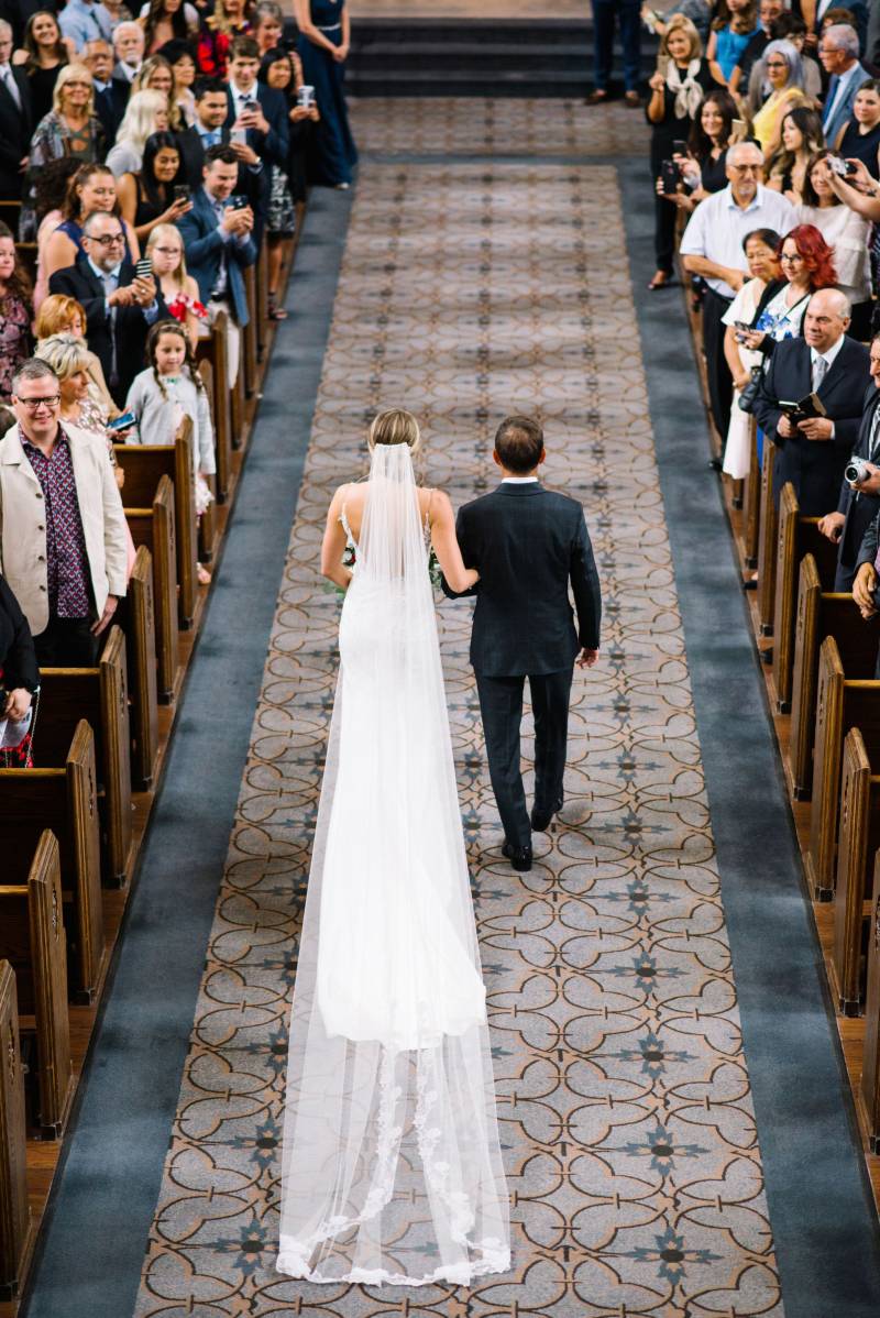 Bride walking the aisle with father with long trailing white veil 