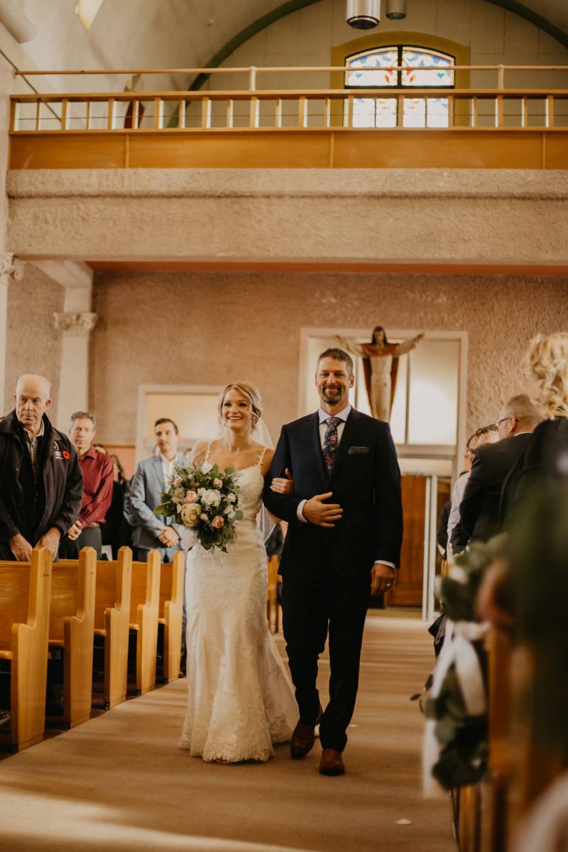 Woman in large white dress holding bouquet walking down church aisle in arms with man in suit