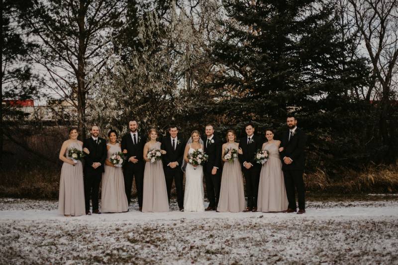 Bride and groom stand beside bridesmaids and groomsmen in line on snowy pathway 