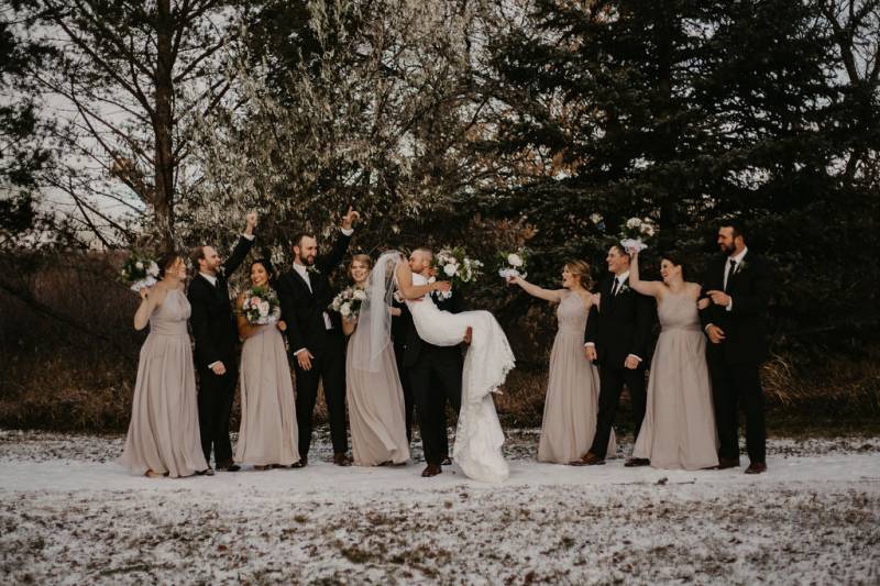 Groom lifting and kissing bride while groomsmen and bridesmaids celebrate behind on snowy pathway
