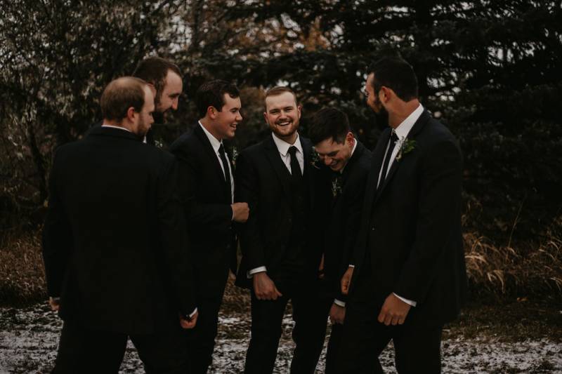 Groom and groomsmen stand together laughing on snowy forest pathway 