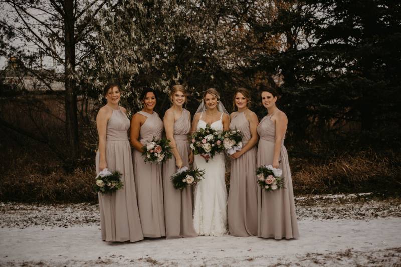 Bride and bridesmaids stand towards holding pink and white bouquets on snowy pathway
