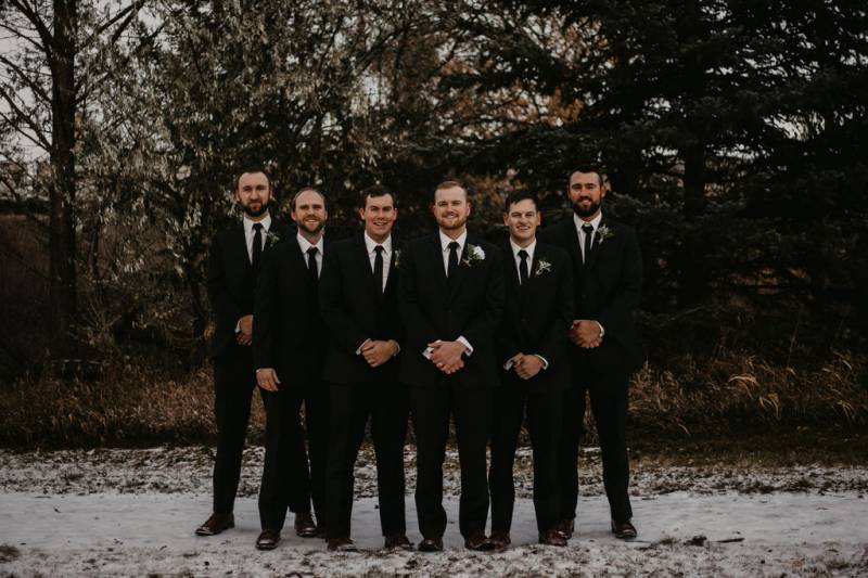 Groom and groomsmen stand crossed arms in line smiling on snowy pathway