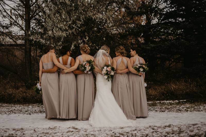 Bride and bridesmaids stand facing away holding waists and bouquets on snowy pathway