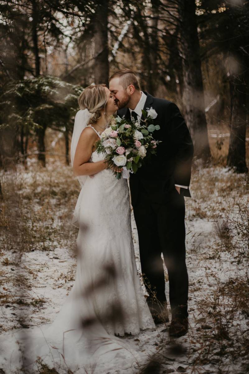 Man and woman in long white dress holding bouquet kiss on snowy pathway