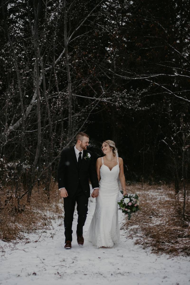 Bride and groom walk holding hands down snowy forest pathway holding pink and white bouquet 