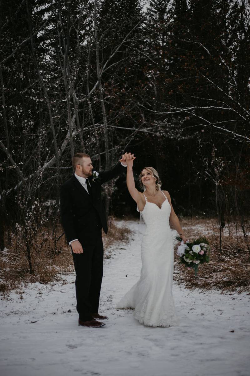 Man and woman wearing white dress hold hands extended over head on snowy forest path