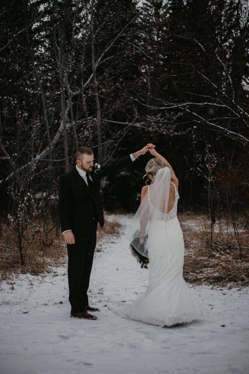 Groom spins Bride in white dress holding bouquet on snowy forest path