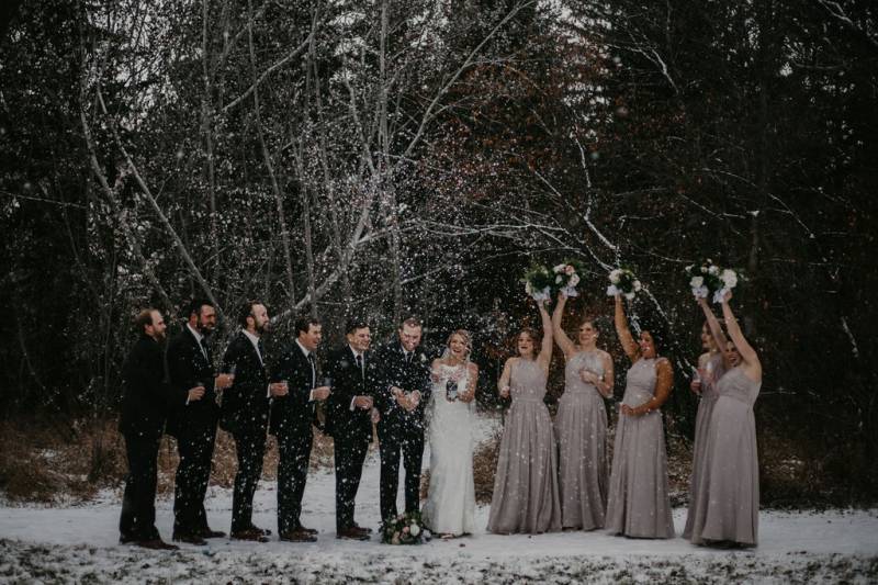 Groom and bride spray champaign while bridesmaids hold white bouquets overhead on snowy forest pathway
