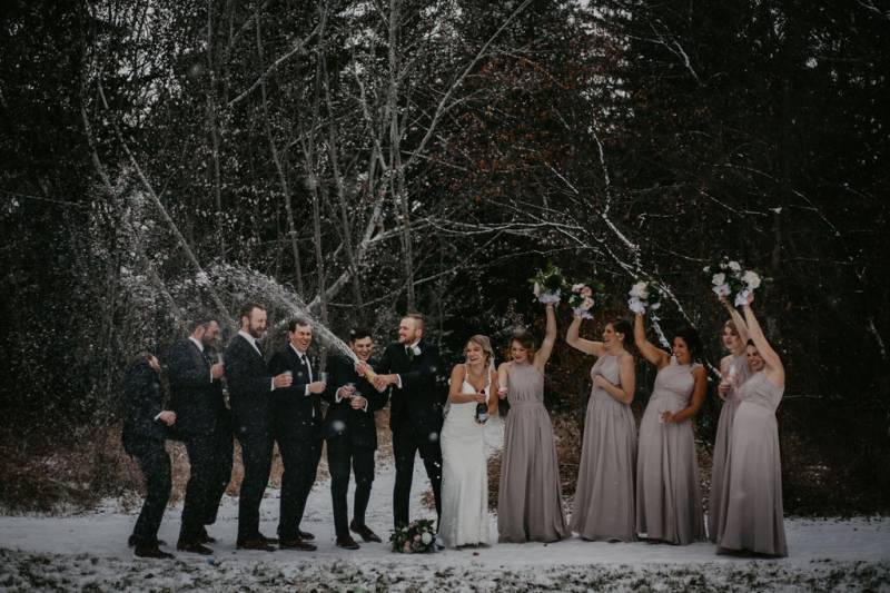 Groom and bride spray champaign while bridesmaids hold white bouquets overhead on snowy forest pathway