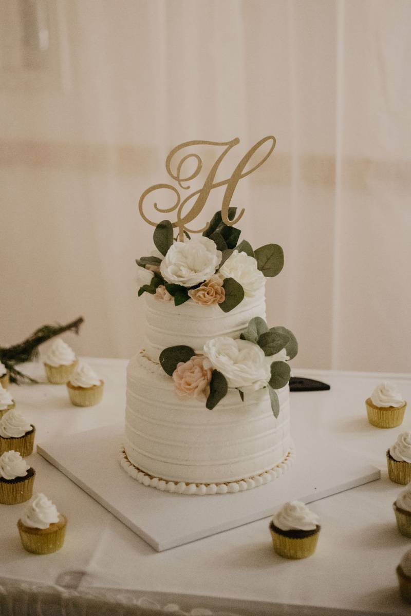 Two tiered white wedding cake with pale pink and white floral accents and gold foil cupcakes