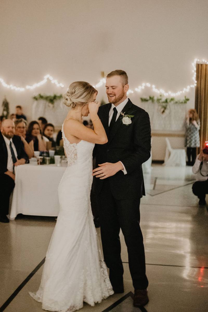 Bride wipes eyes standing in white dress and groom smiles 