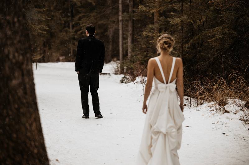 Bride in white dress stands away from man in black suit on snowy path