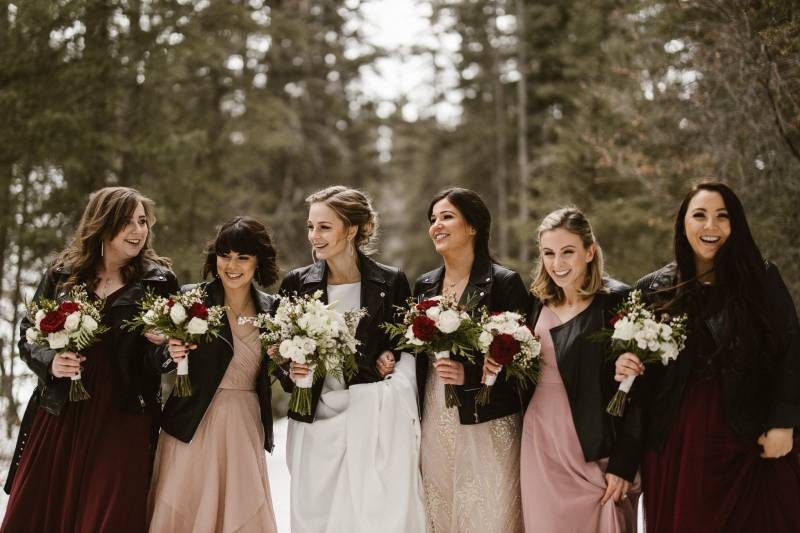 Bride and bridesmaids walk arms linked holding burgundy and white bouquets 
