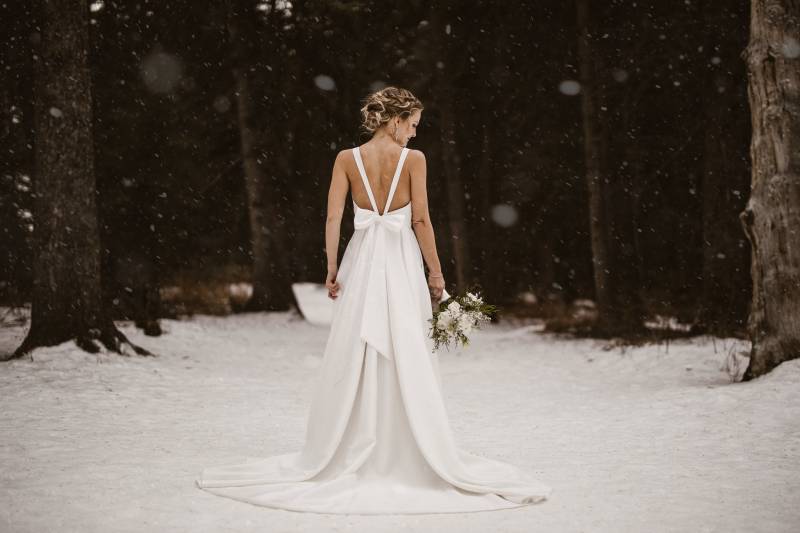 Bride standing in white dress holding white bouquet to side in snowy forest field