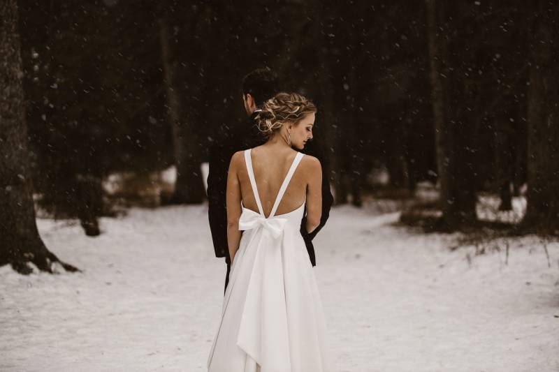 Bride stands behind groom looking back in snowy forest clearing 