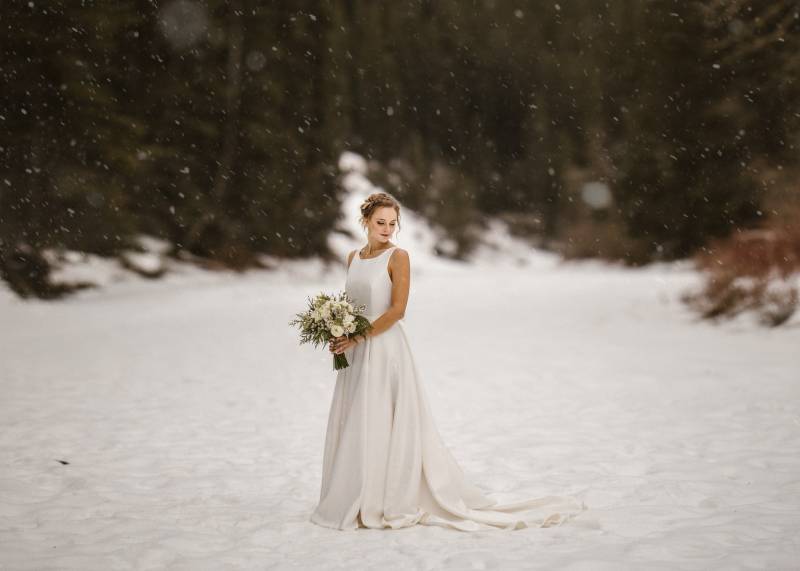 Bride in white dress holding white bouquet standing in snowy field 