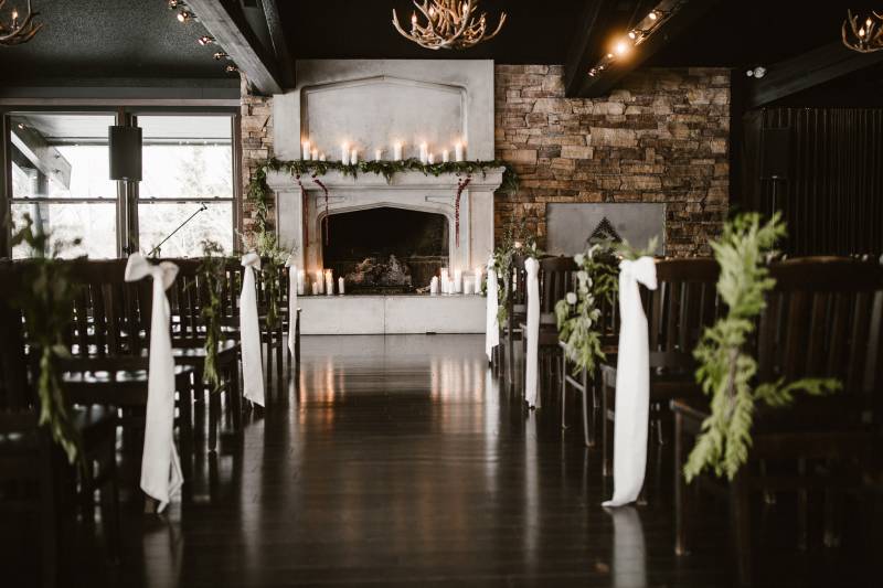 Candle lit wedding reception with black benches with white bows facing large white fireplace 