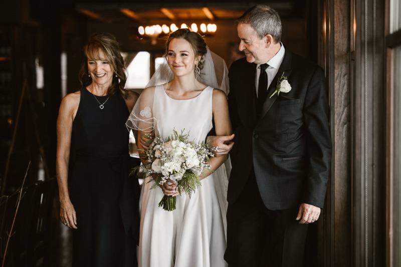 Bride walks with man and woman smiling while linking arms 