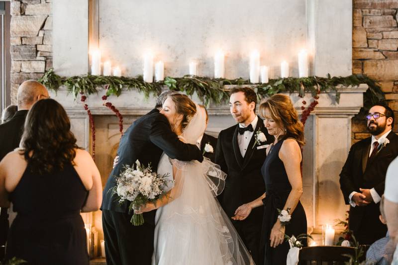 Bride in white dress and veil embraced by man in black suit by fireplace
