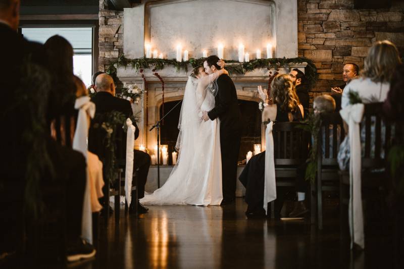 Bride and groom kiss while embracing by candle lit fireplace 