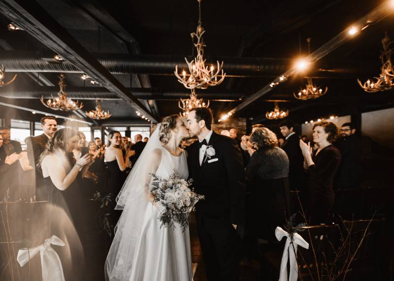 Bride and groom kiss underneath antler chandeliers as guests applaud