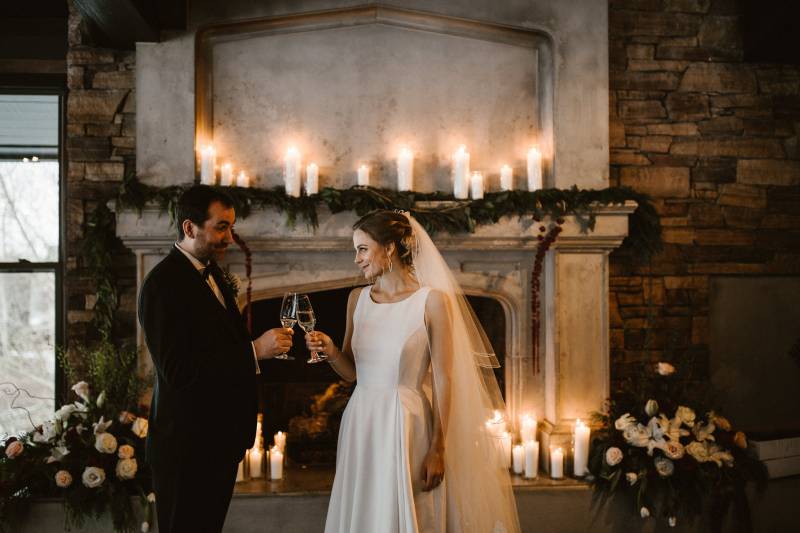 Bride and groom clink glasses in front of candle lit fireplace 