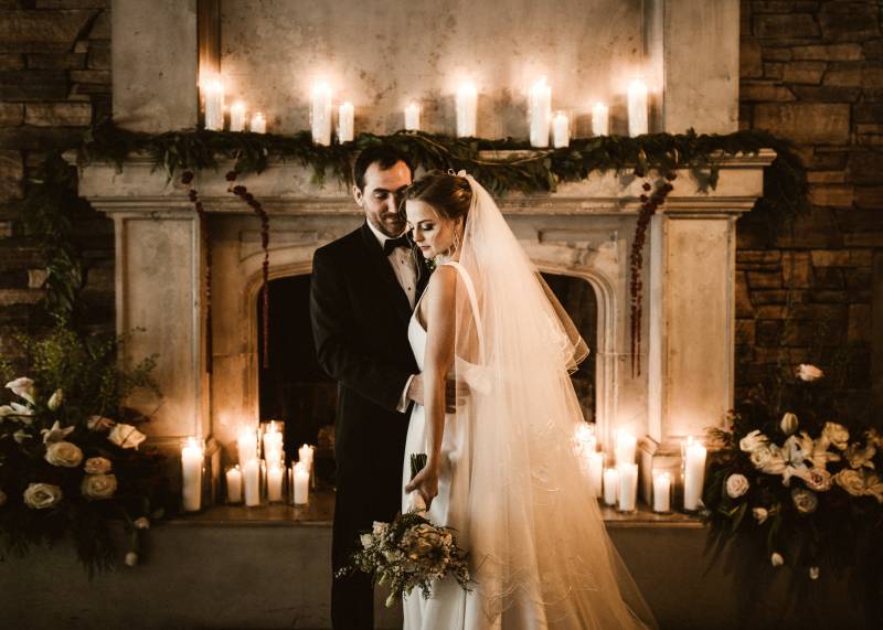 Groom holding bride waist holding white bouquet in front of candle lit fireplace 
