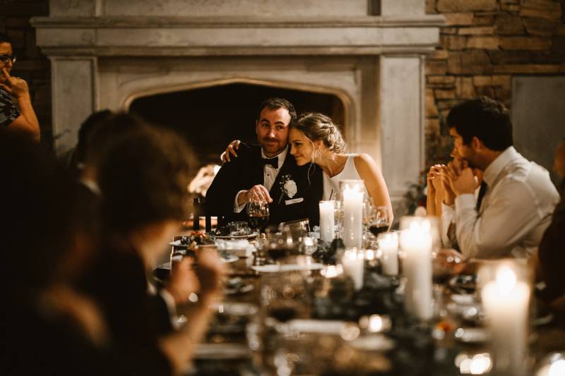Bride and groom embrace at the end of candle lit table in front of fireplace
