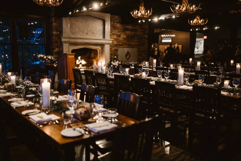 Black wood chairs seated around long brown tables under dimly lit candles and antler chandeliers 