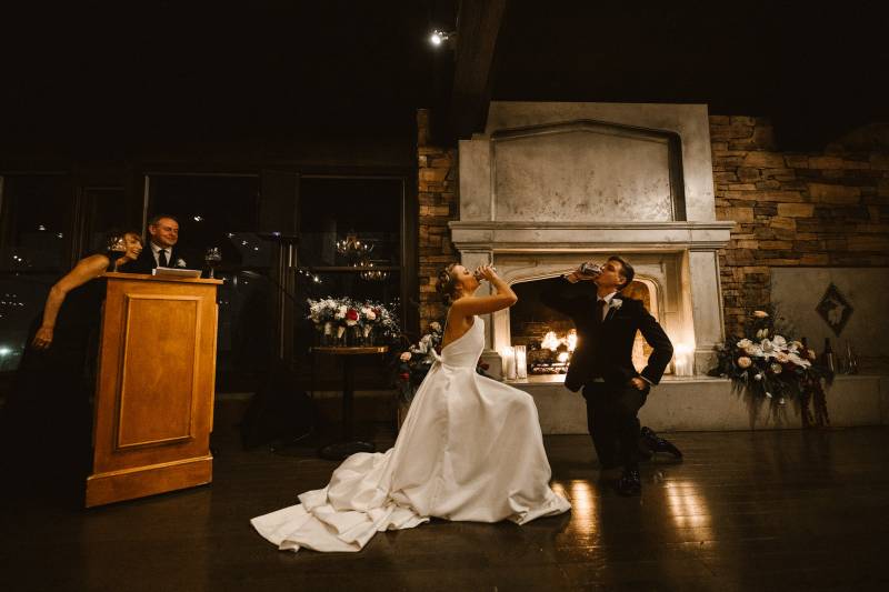 Bride and groom drink from can on one knee in front of fireplace and podium 