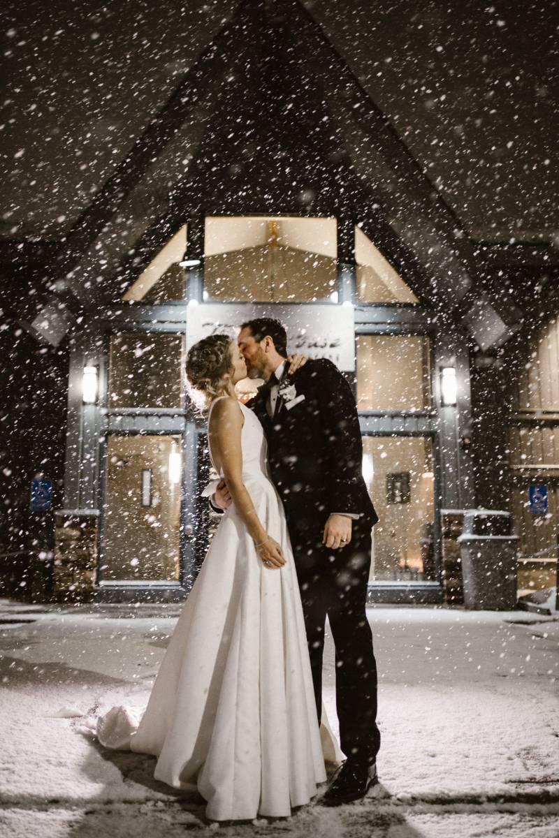 Bride and groom kiss outside large black cabin while snow falls around them 