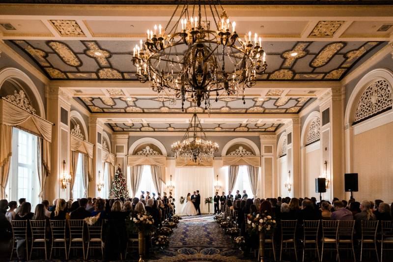 Wedding ceremony in large cream and gold room with hanging chandeliers 