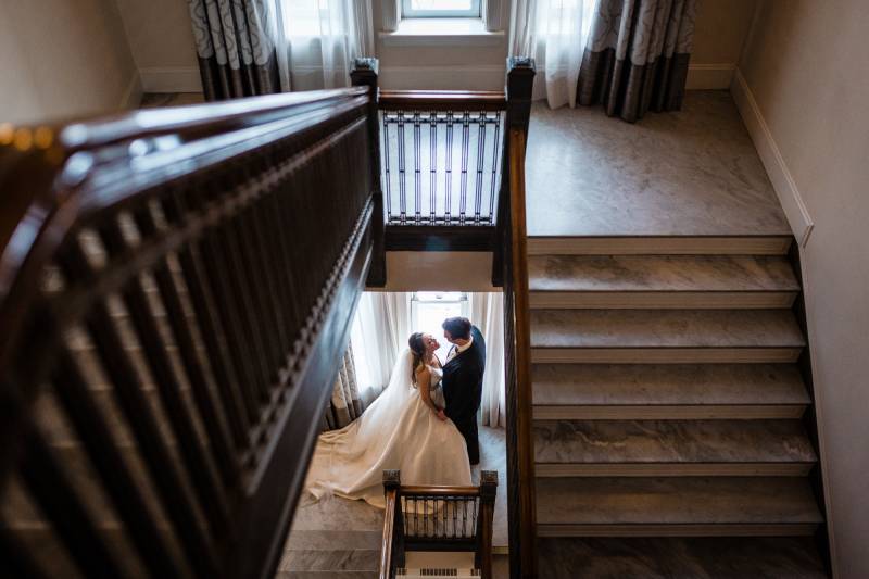 Bride and groom stand embraced at bottom of winding staircase 